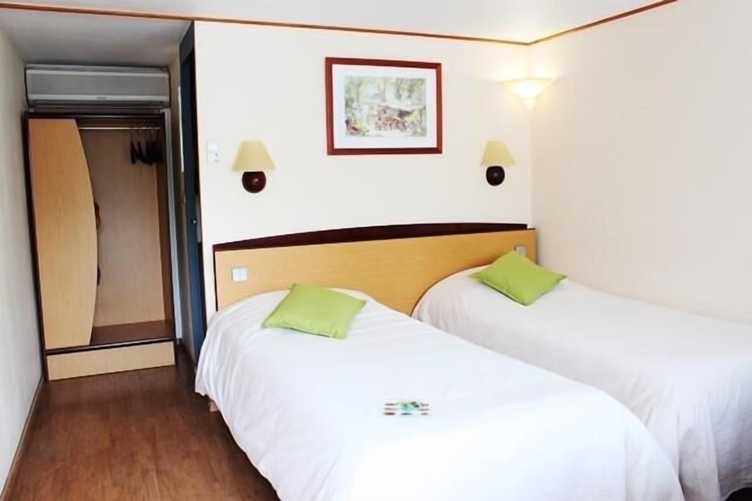 Een hotelkamer met twee enkele bedden, een houten garderobe, een ingelijst schilderij aan de muur en twee wandlampen.