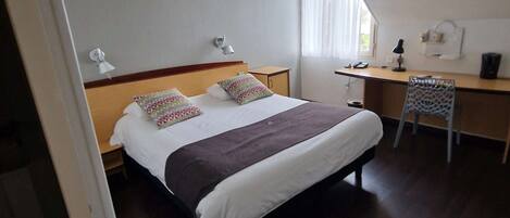 A hotel room with a bed, a desk, and a chair. There is a window with curtains, a lamp on the desk, and a wall-mounted phone.