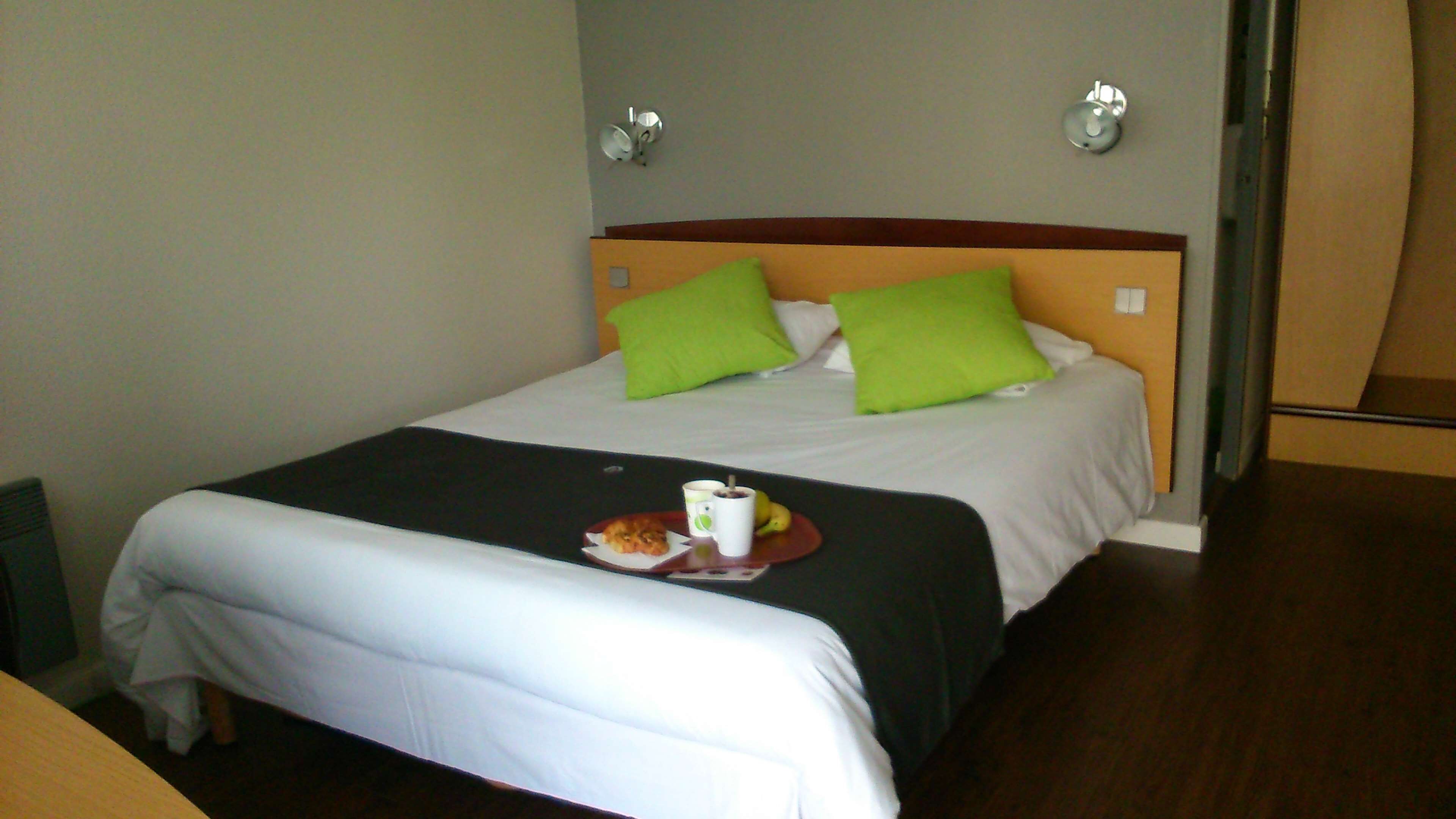 A hotel room with a bed, green pillows, a bedside table with a plate of food and a cup, and a wall-mounted light fixture.