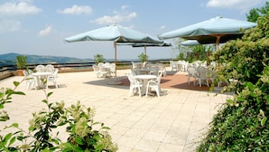 Un patio al aire libre con mesas y sillas de plástico blanco, cubierto por grandes sombrillas, rodeado de follaje.