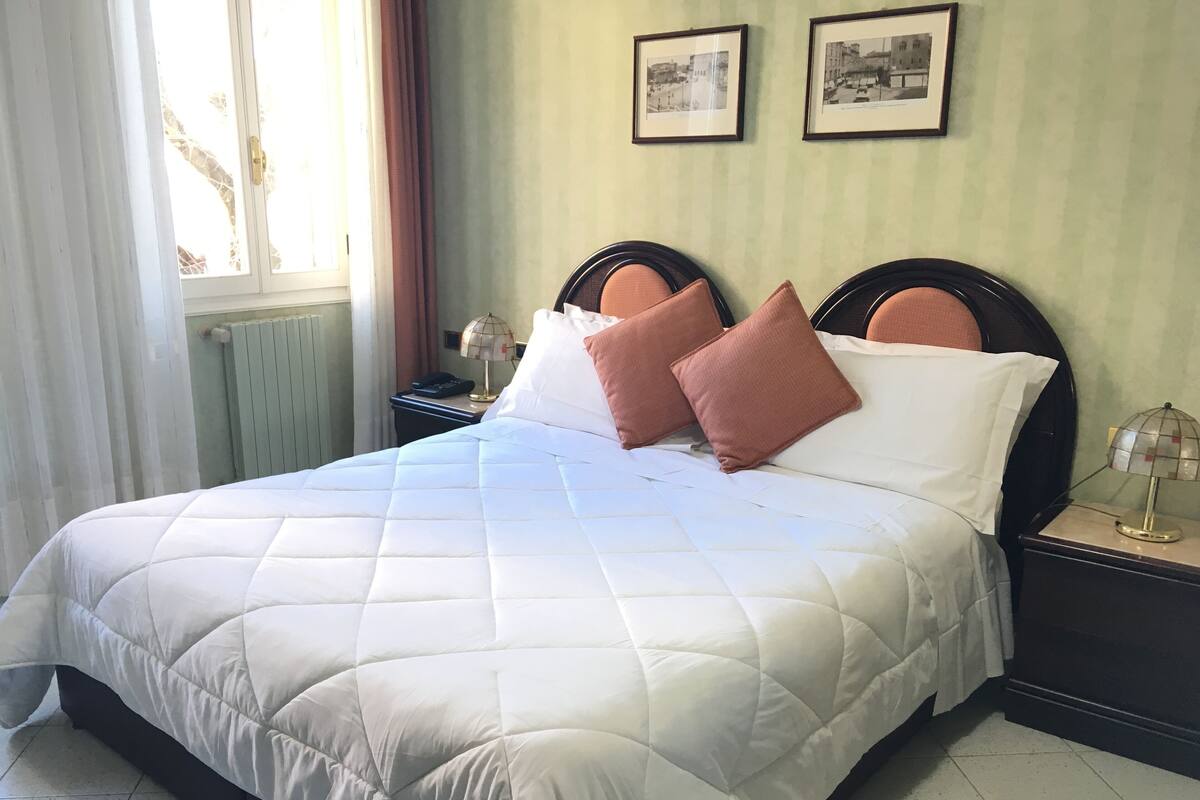 A neatly made bed with a white quilted bedspread and two pink pillows, a bedside table with a lamp, and two framed pictures on the wall.