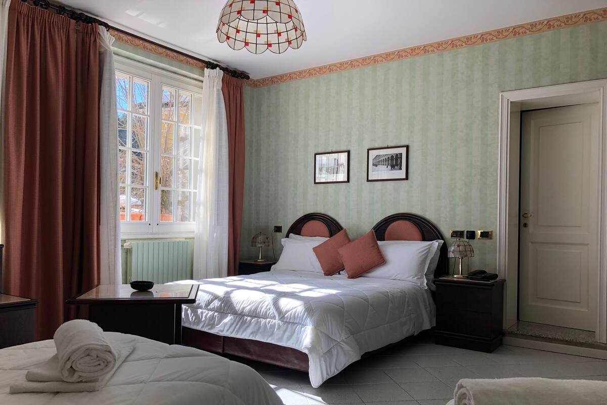 A hotel room with a bed, two bedside tables, a window with curtains, a door, and two framed pictures on the wall.