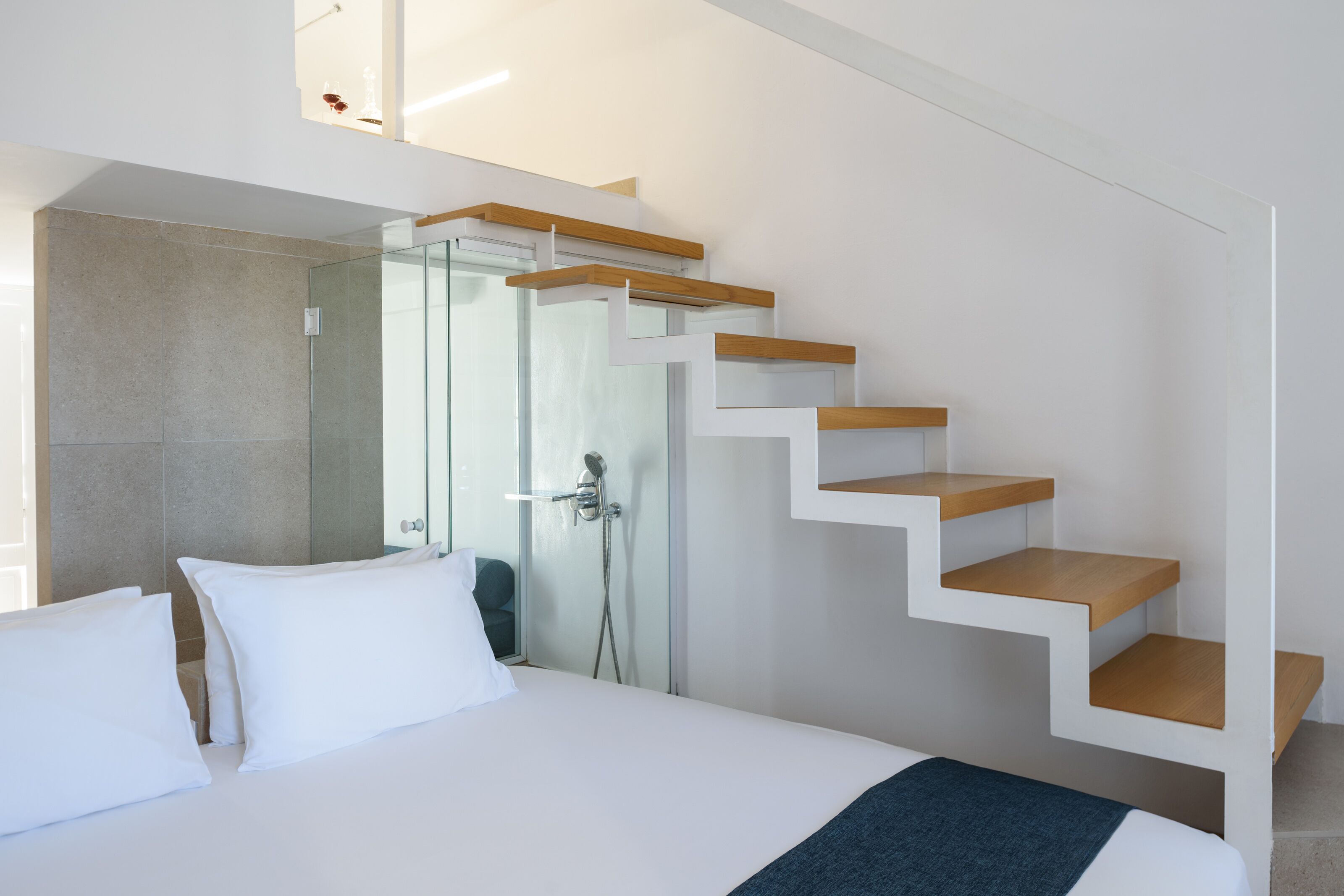 A modern hotel room with a bed, white pillows, a blue throw blanket, a glass shower enclosure, and a wooden staircase.