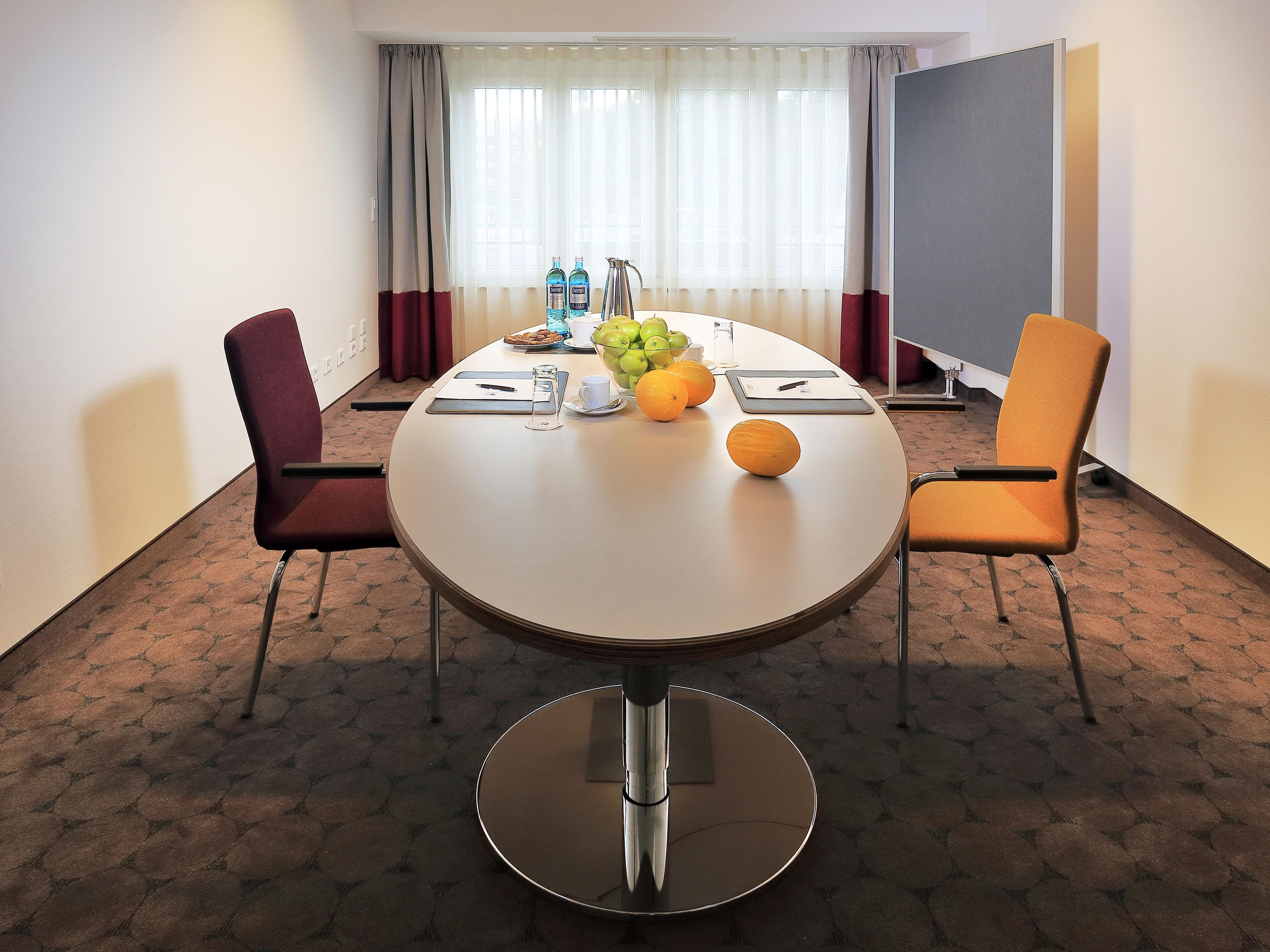 Uma sala de reunião de hotel com uma mesa redonda, duas cadeiras, uma cesta de frutas e um dispensador de água.