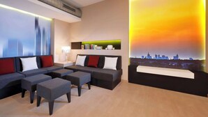 A modern hotel room with a grey sofa set, matching ottomans, a cityscape mural, and a small shelf with books and decorative items. - Best Western Premier IB Hotel Friedberger Warte (Frankfurt)