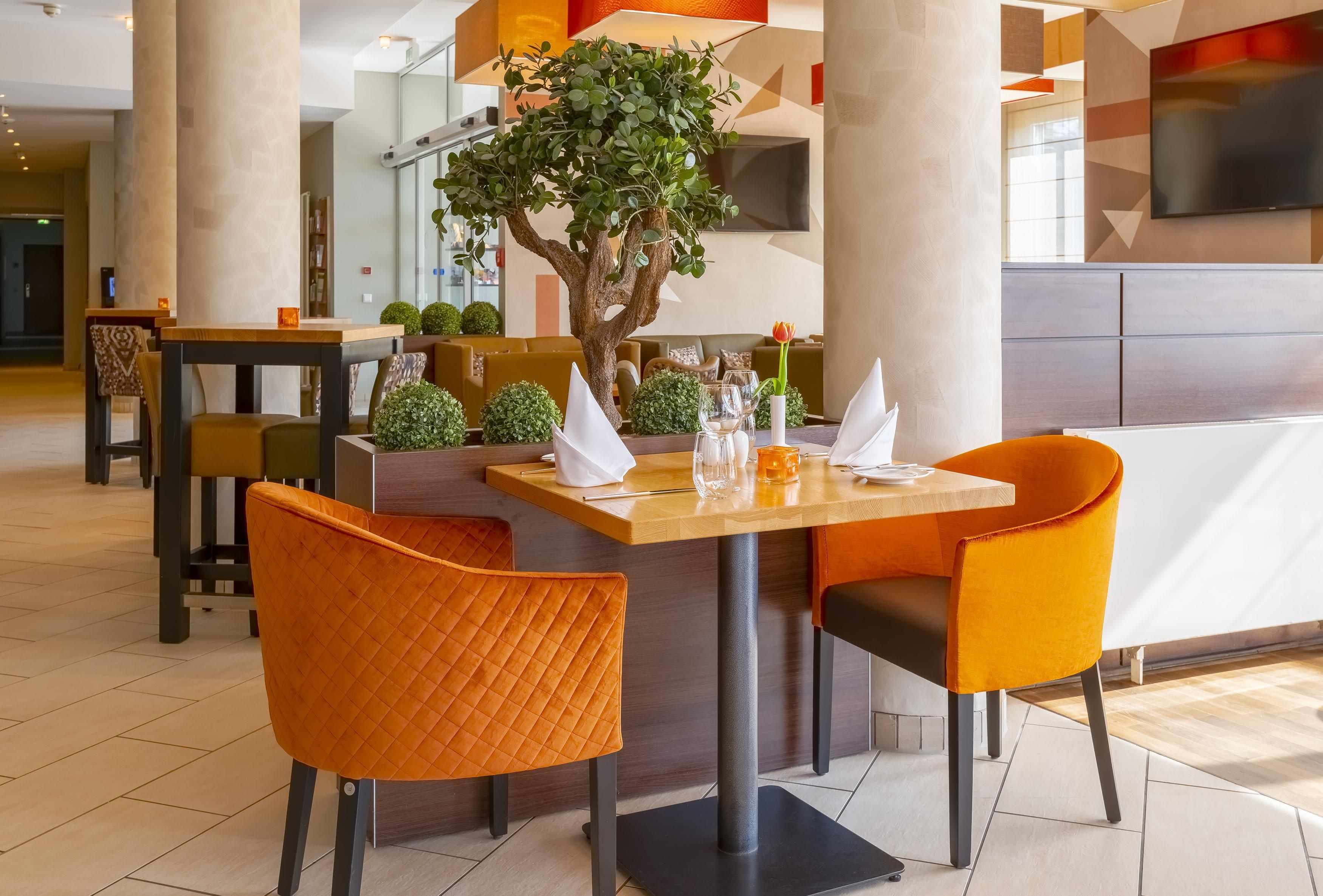 Comedor con mesa dispuesta para cuatro personas, sillas naranjas, un árbol en maceta y diseño de interiores moderno.