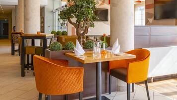 Comedor con mesa dispuesta para cuatro personas, sillas naranjas, un árbol en maceta y diseño de interiores moderno.