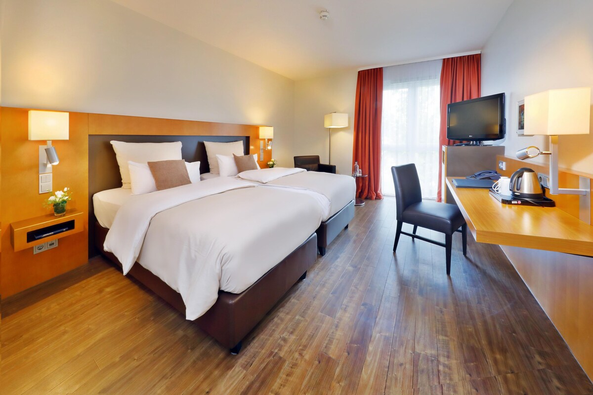 A hotel room with a double bed, bedside tables, a desk with a chair, a television, and a window with curtains.