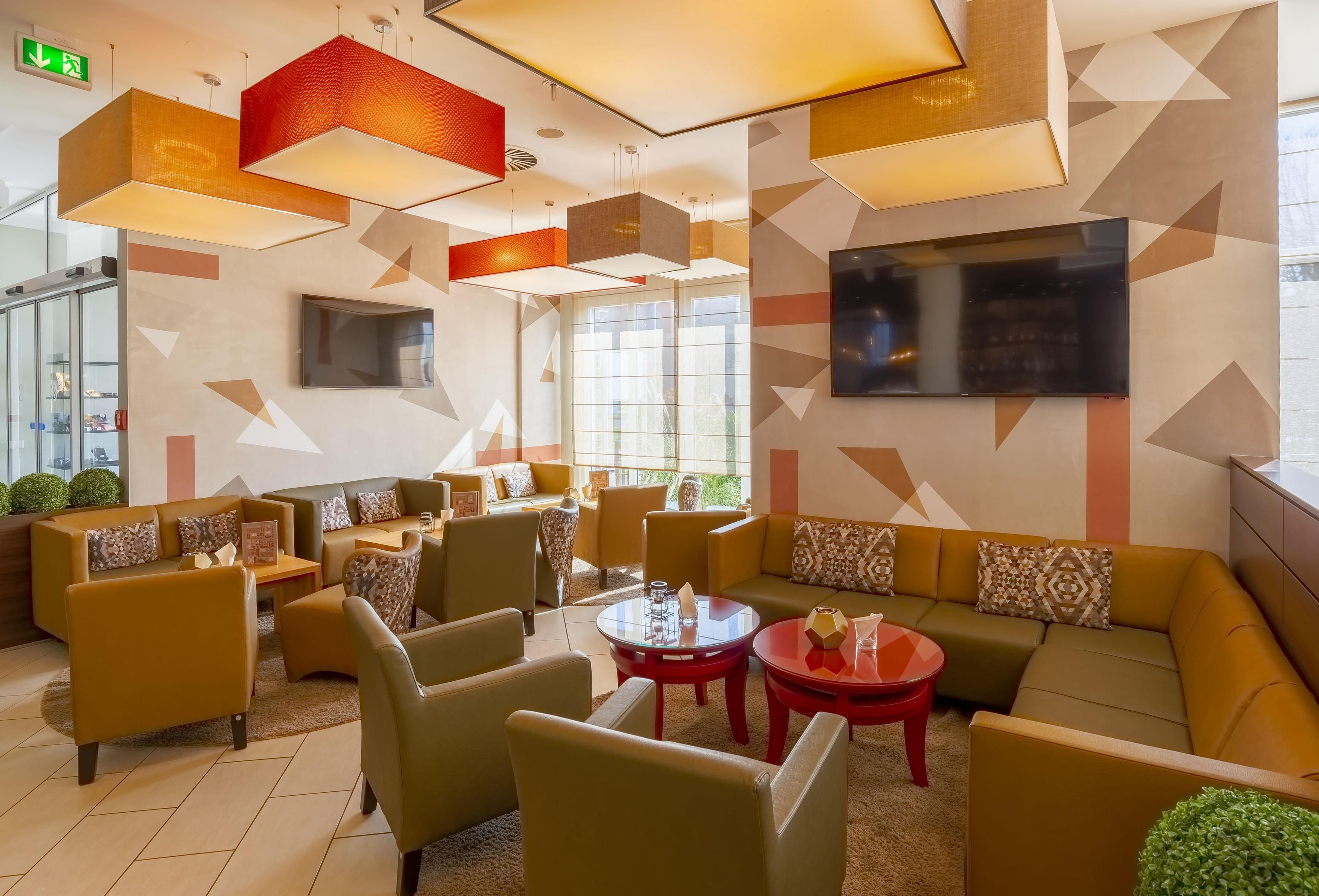 A modern lounge area with sofas, chairs, and tables, featuring geometric wall patterns and a flat-screen TV.