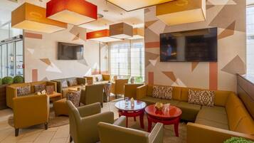 A modern lounge area with sofas, chairs, and tables, featuring geometric wall patterns and a flat-screen TV.