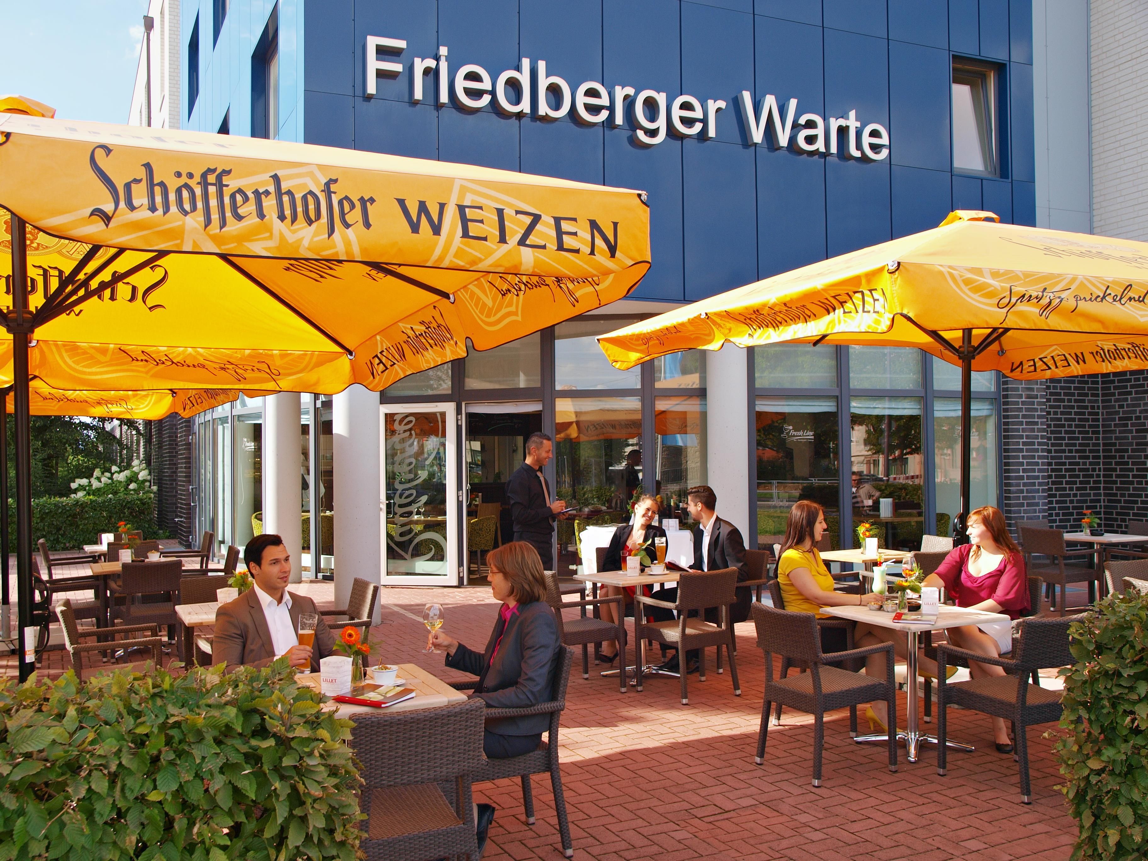 Ein Außenbereich mit Tischen und Stühlen unter gelben Sonnenschirmen, vor einem Gebäude mit der Aufschrift 'Friedberger Warte'.