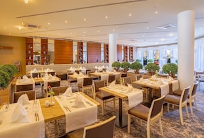 A hotel dining area with multiple tables set for guests, featuring a modern interior design with wooden accents and a carpeted floor.
