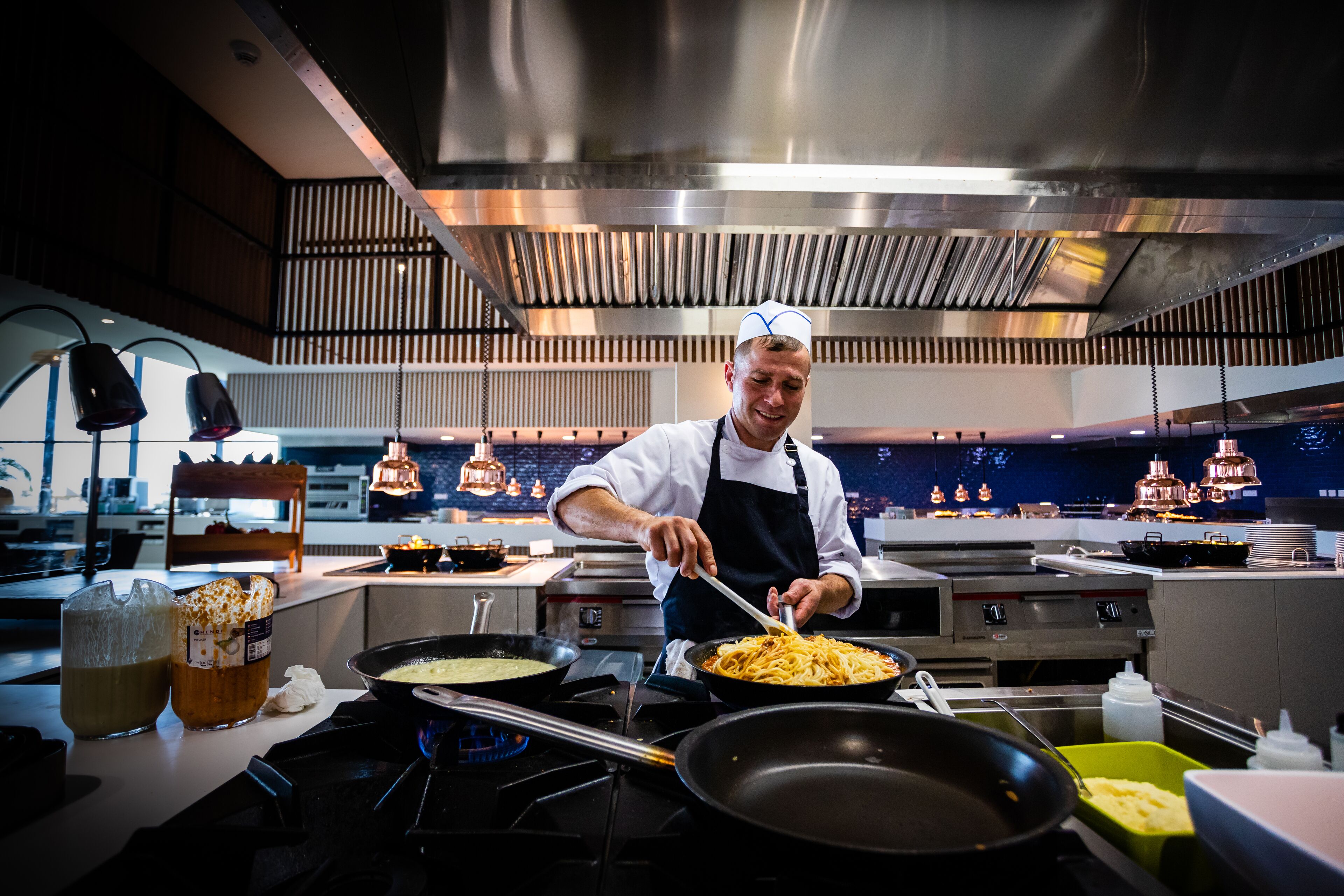 A chef cooking in a modern kitchen with stainless steel appliances and a high ceiling.