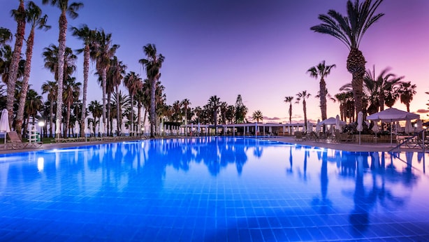 Una piscina con sillas de descanso y sombrillas, rodeada de palmeras.