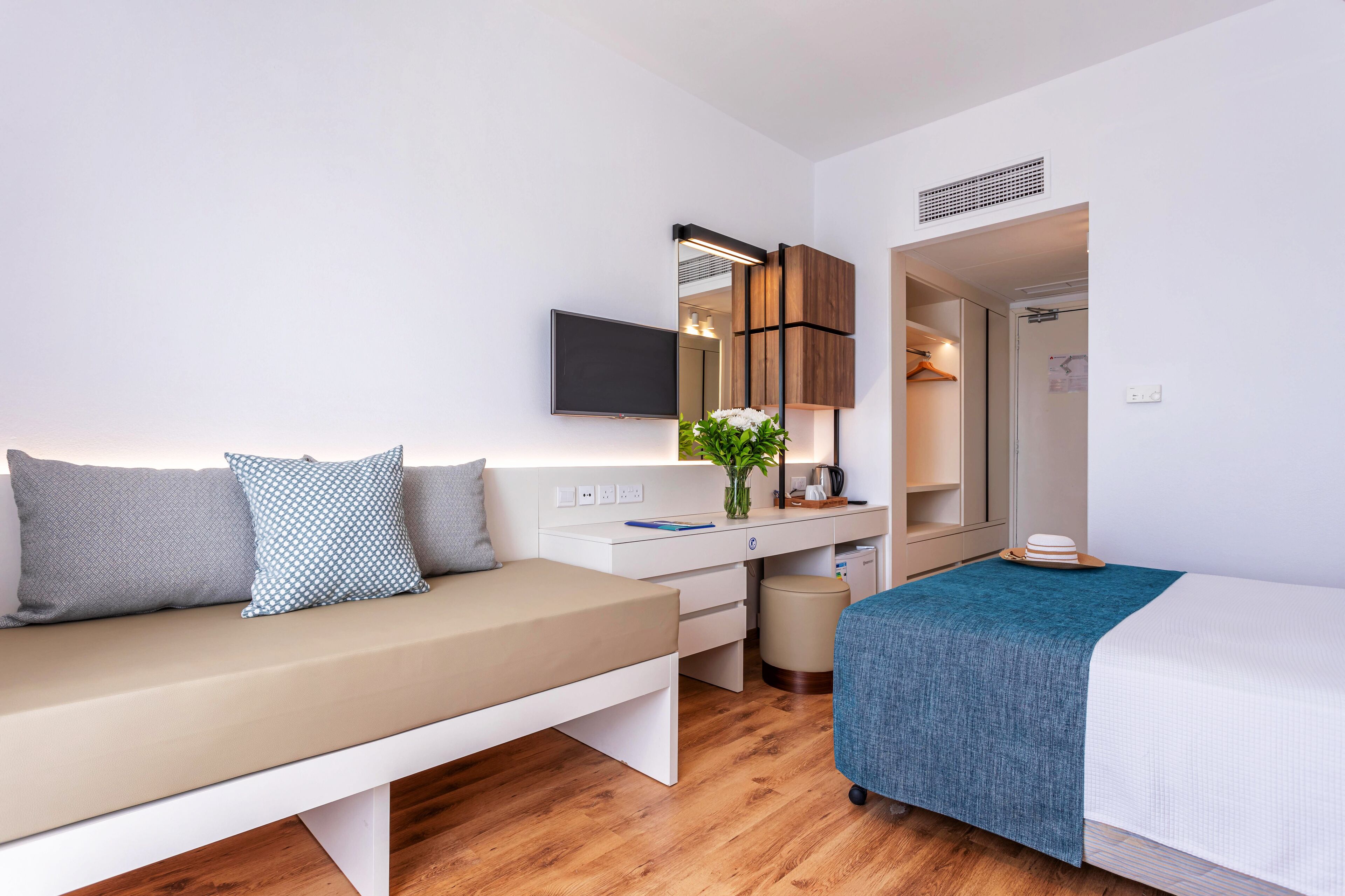 A modern hotel room with a sofa, a bed, a desk with a mirror, a television, and a small bathroom.
