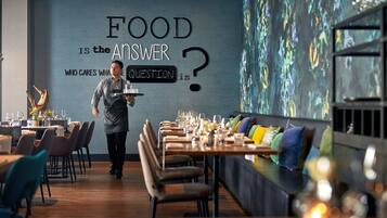 A restaurant interior with a waiter carrying a tray. Tables are set with wine glasses and cutlery. A wall features a quote about food.