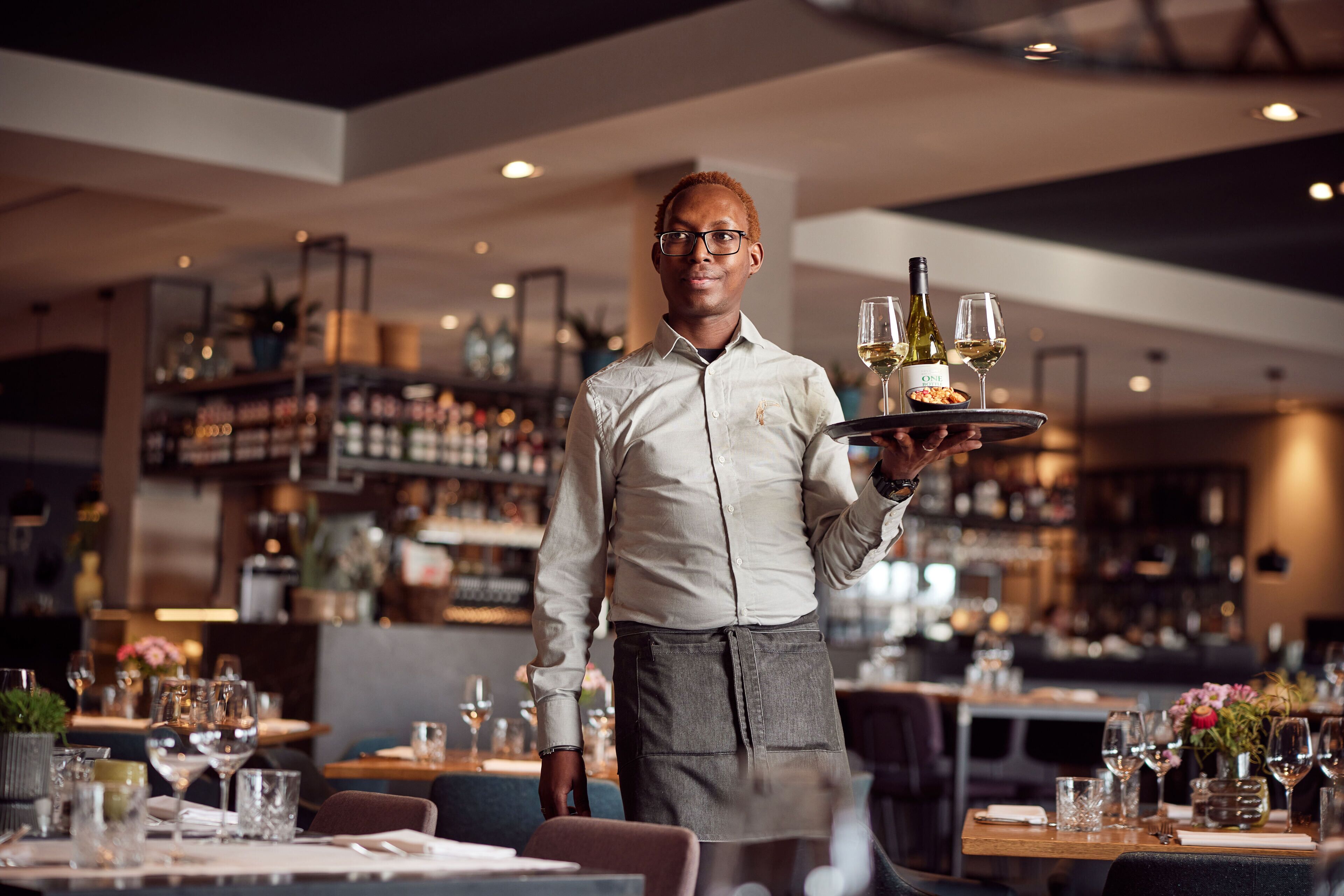A waiter in a restaurant holding a tray with wine glasses and a bottle.