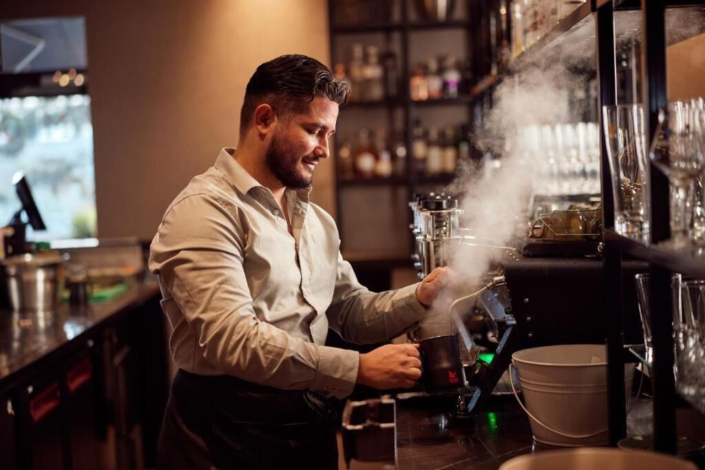 Una persona fent servir una màquina de cafè en un bar, amb estantes plenes de botelles i gots al fons.