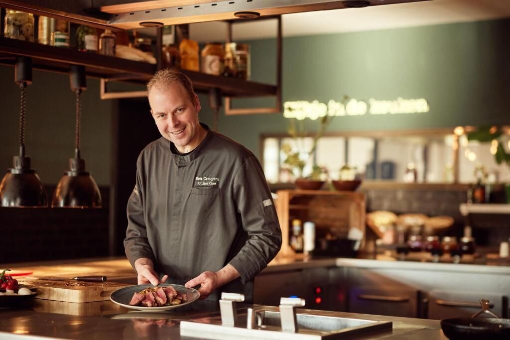 Een kok in een keuken met een houten aanrecht, hangende lampen en planken met potten.