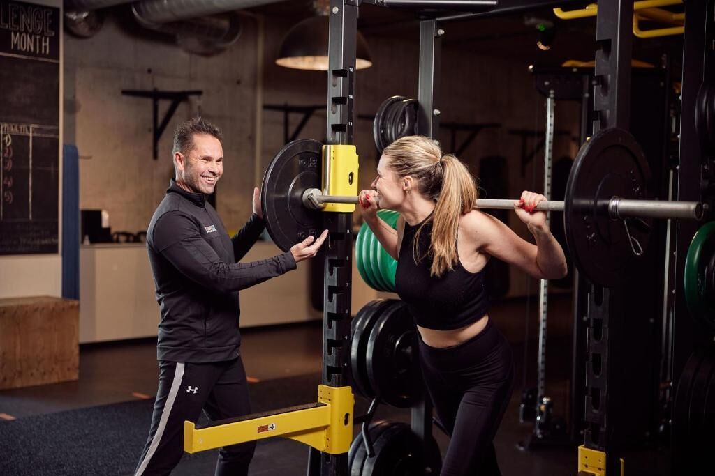 A gym with weightlifting equipment, a chalkboard, and a person assisting another person with a weightlifting exercise.