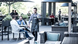 An outdoor dining area with wicker furniture, umbrellas, and a waiter serving a table. - Van Der Valk Hotel Stein - Urmond (Urmond)
