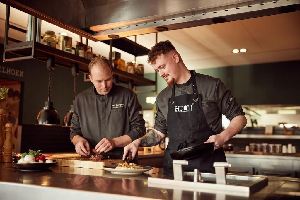 Two chefs in a kitchen with stainless steel appliances and wooden shelves.