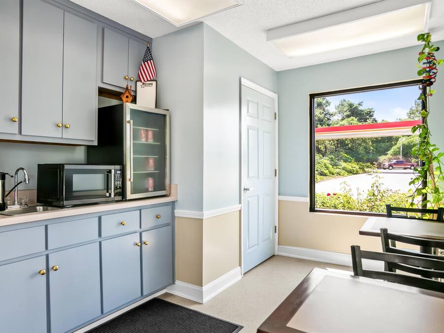A compact kitchen with grey cabinets, a microwave, and a small refrigerator. There's a window with a view of a parking lot and greenery.