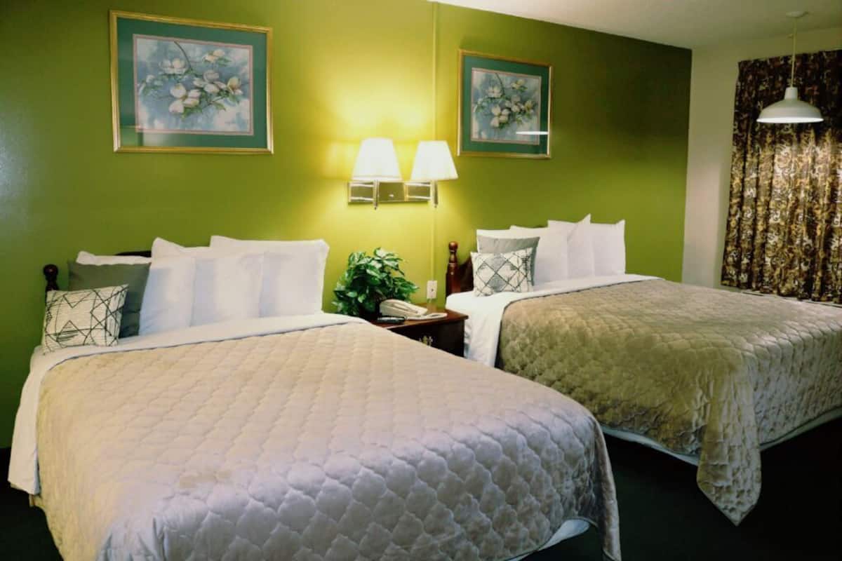 Ein Hotelzimmer mit zwei Betten, grünen Wänden, floralen Bildern, einem Nachttisch mit Lampe und einem Vorhang.