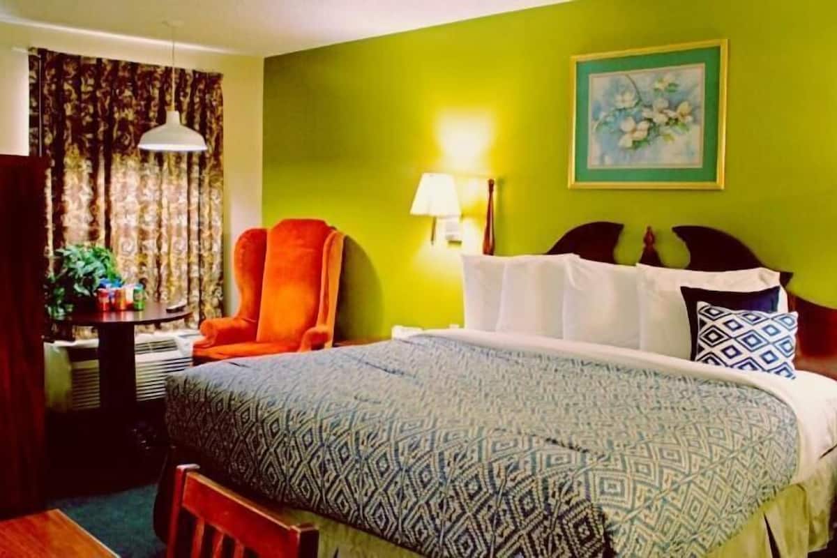 Ein Hotelzimmer mit Bett, Sessel, Tisch mit Lampe, einem Bild an der Wand und Vorhängen.