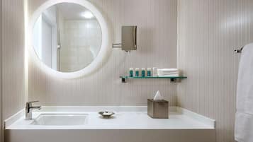 Une salle de bain moderne avec un grand miroir rond, un lavabo blanc et une étagère en verre où sont rangés les articles de toilette.