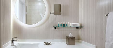 A modern bathroom with a large round mirror, a white sink, and a glass shelf with toiletries.