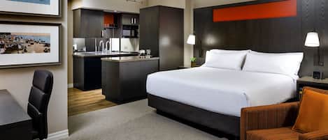 A hotel room with a large bed, a desk, a chair, a small table, a kitchenette, and framed pictures on the wall.