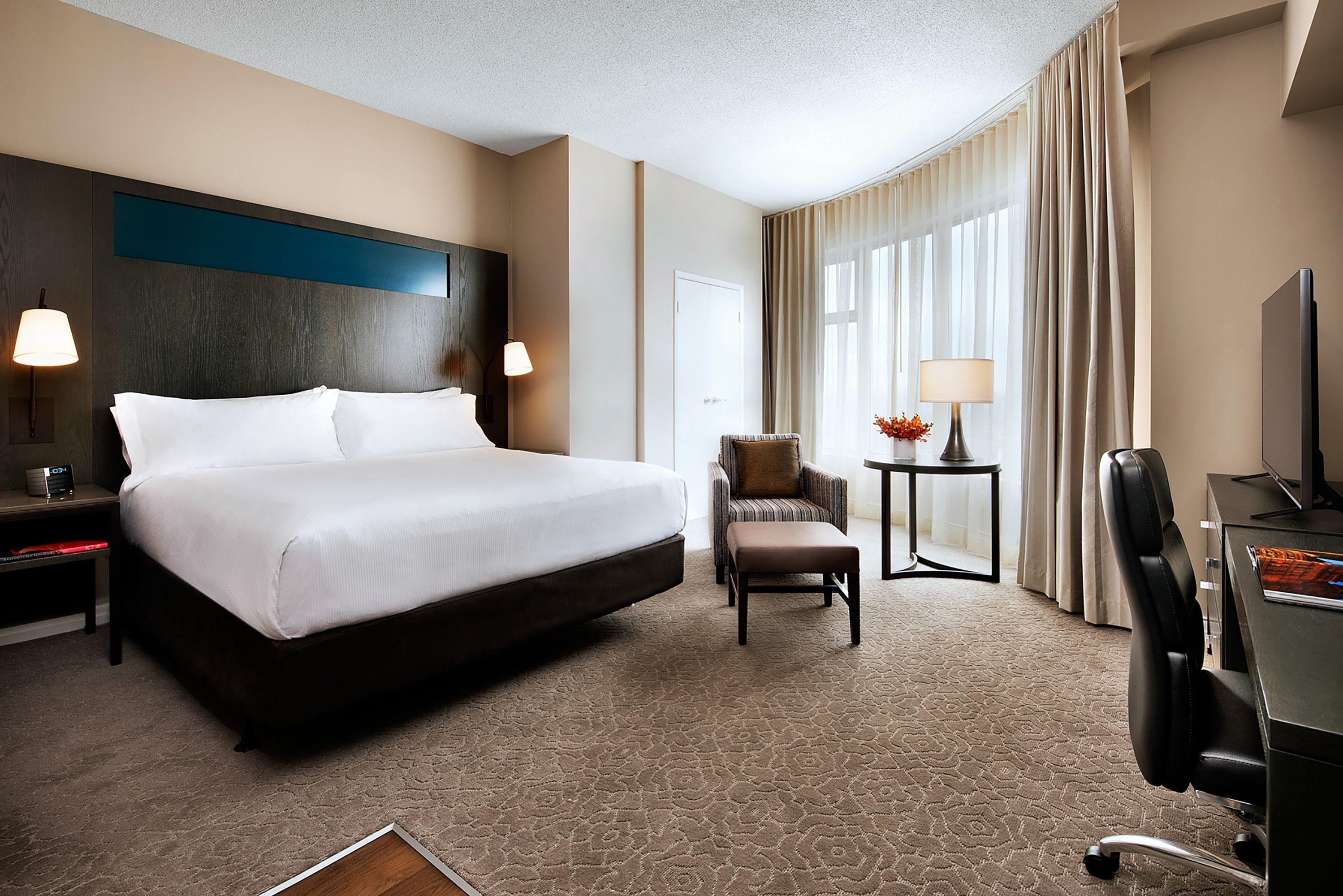 A hotel room with a large bed, a chair, a desk, a lamp, and a TV.