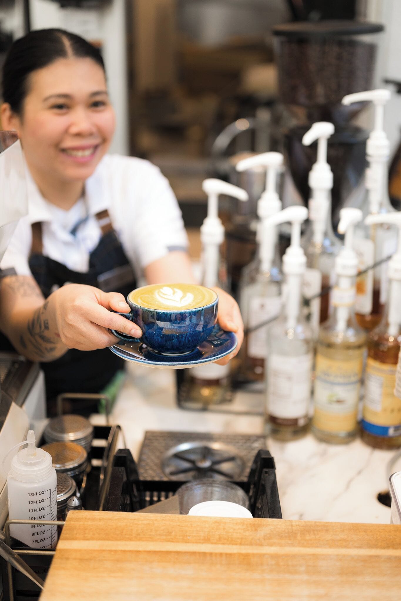Un barista sostiene una taza de café con arte latte, con equipo para preparar café y botellas al fondo.