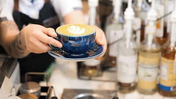 Un barista sostiene una taza de café con arte latte, con equipo para preparar café y botellas al fondo.