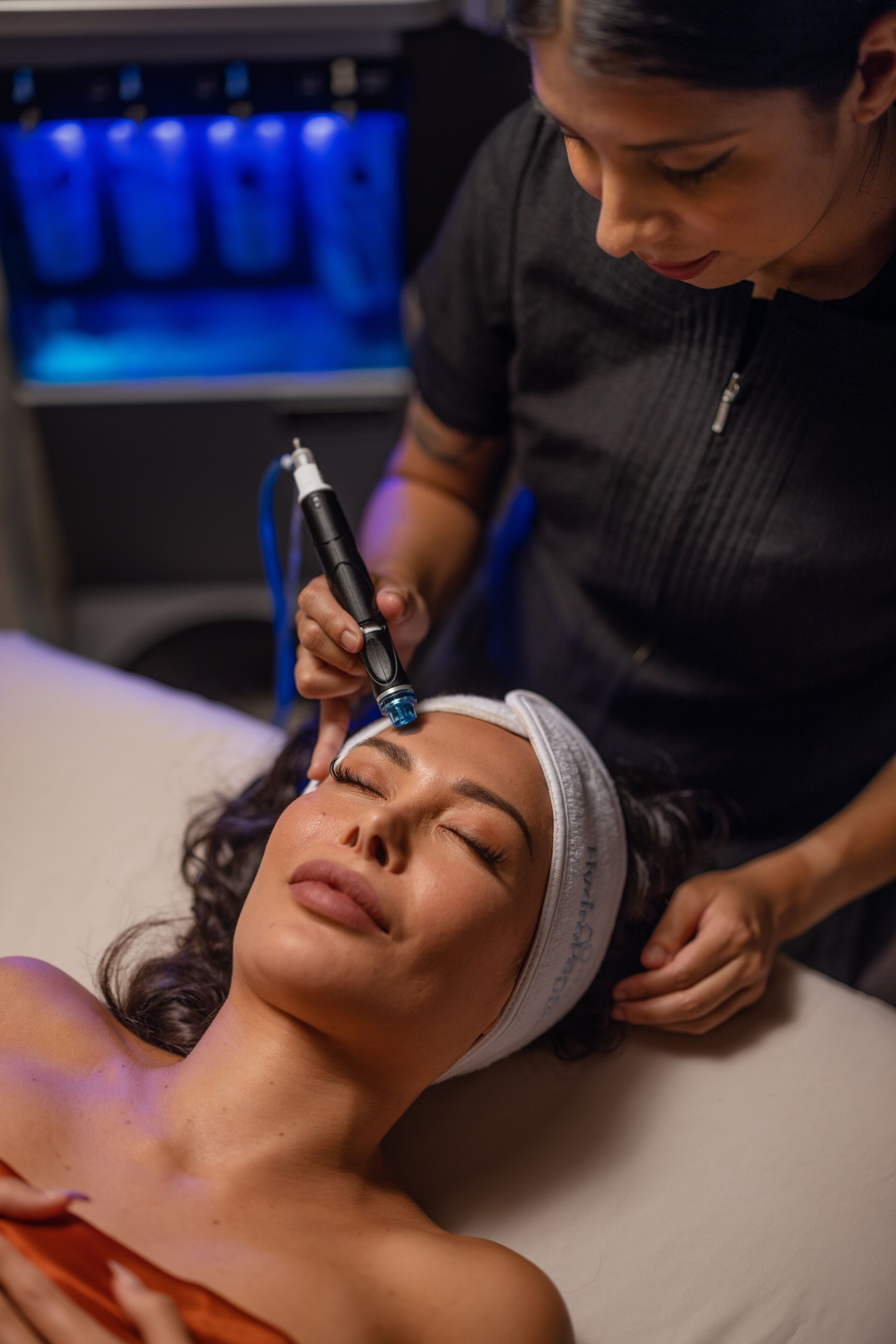 A person lying on a bed with a headband, another person holding a medical device, and a blue-lit machine in the background.