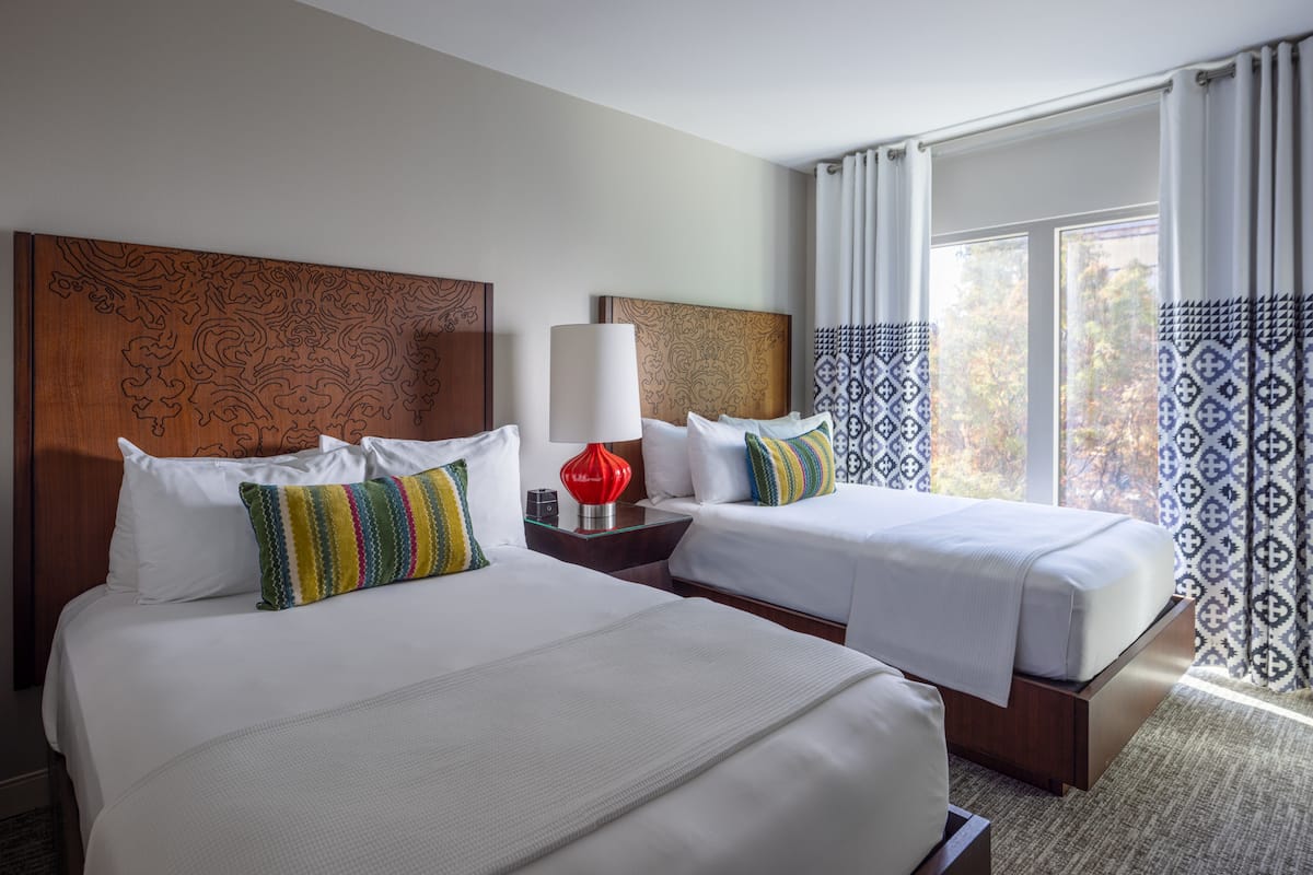 Une chambre d’hôtel avec deux lits, une lampe rouge, des rideaux à motifs et une vue sur les arbres par la fenêtre.
