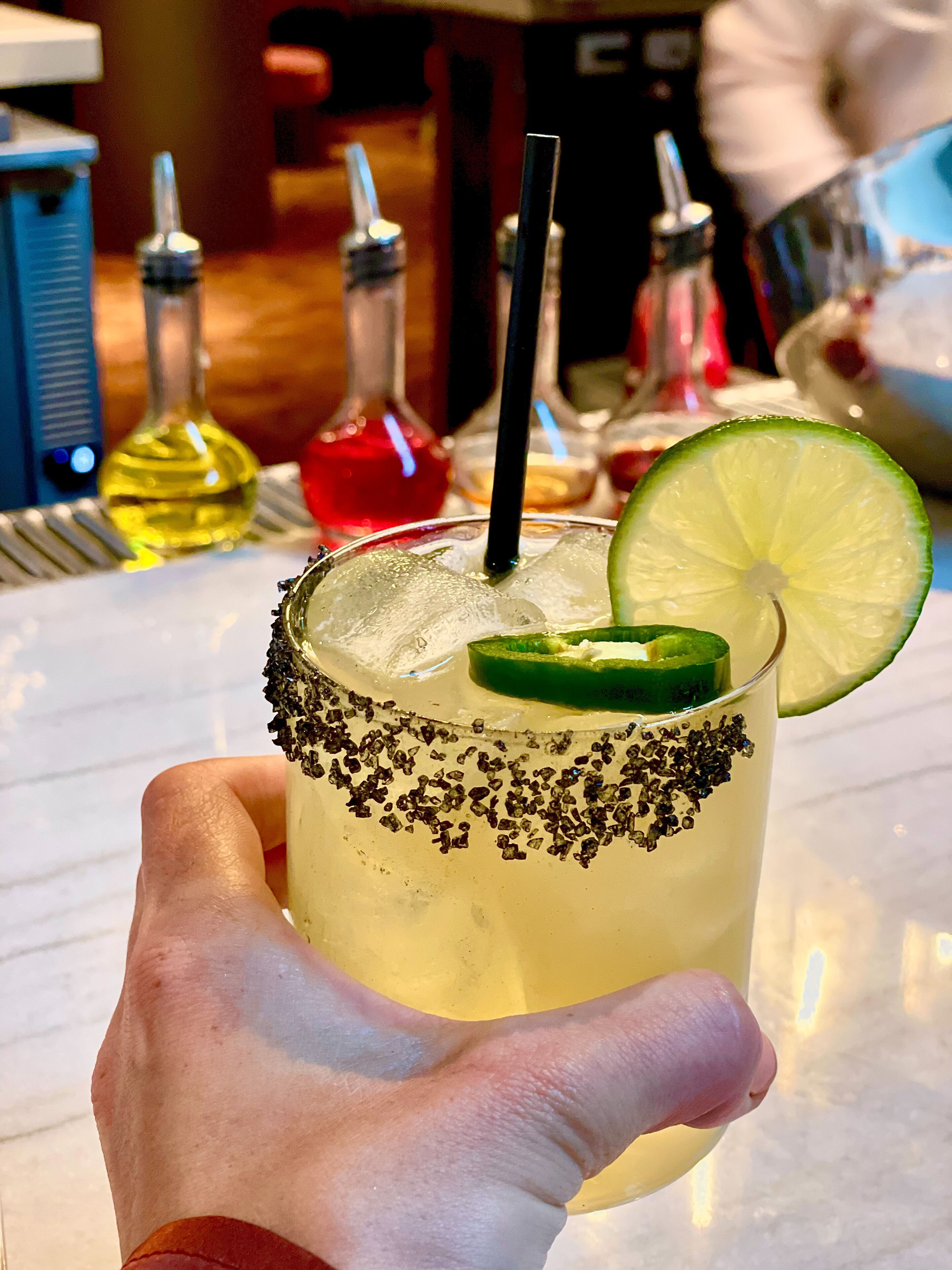 Una mano sosteniendo una margarita con borde de sal, rodaja de lima y pajita negra.