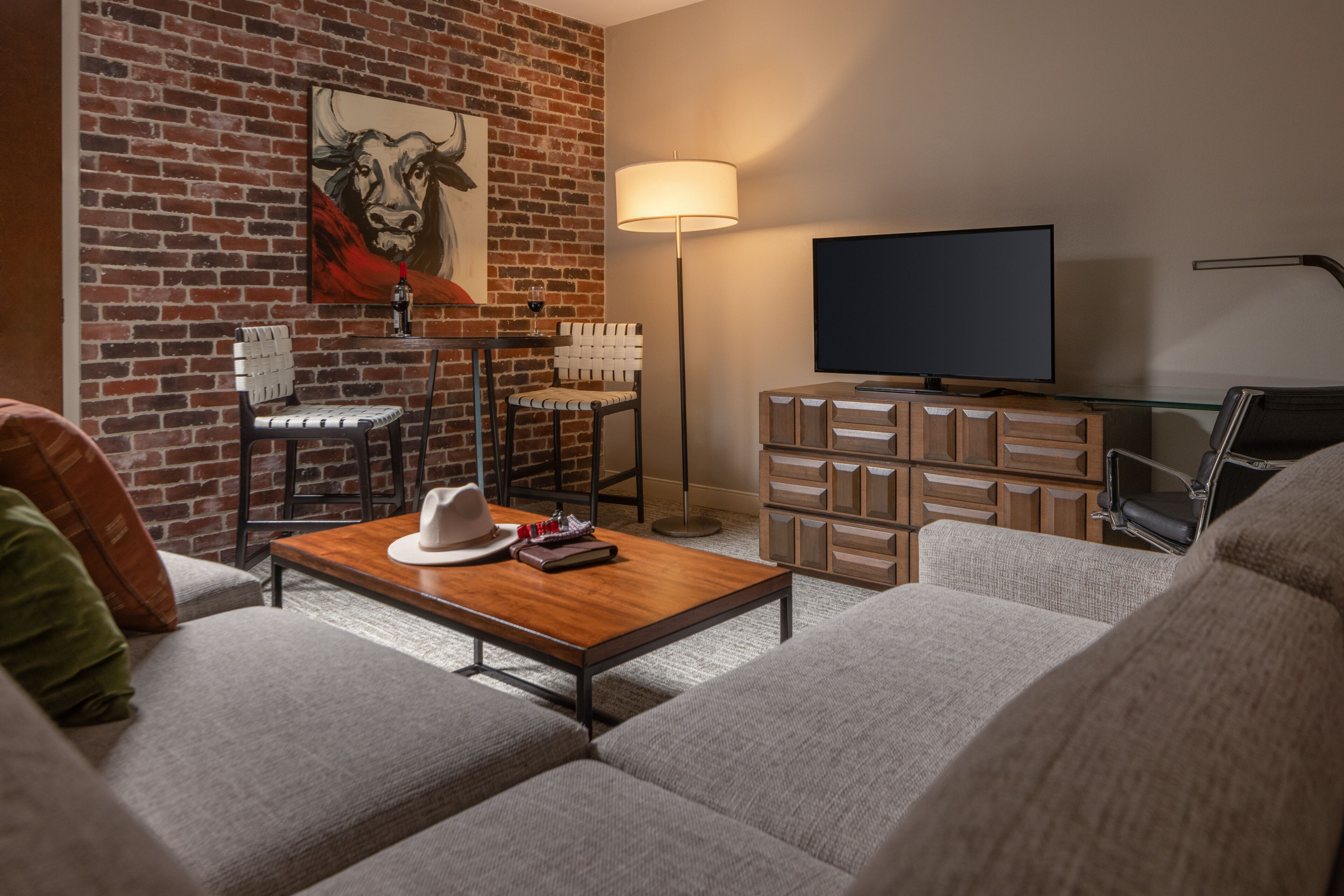A modern living room with a brick wall, a flat-screen TV, a wooden coffee table, a sofa, and a lamp.