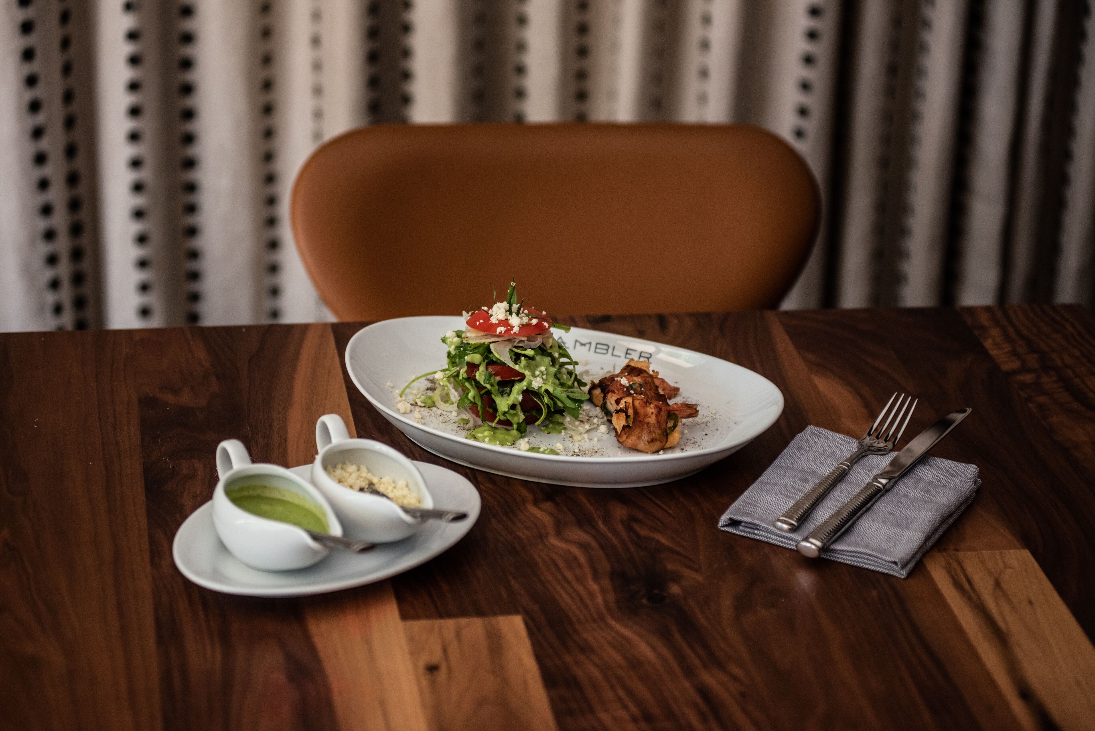 A wooden table with a plate of food, a bowl of soup, and a napkin with cutlery.
