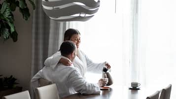 Un couple est assis à une table devant une machine à café et une tasse, dans une pièce où se trouvent une lampe moderne et une plante.