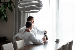 Una pareja sentada en una mesa con una cafetera y una taza, en una habitación con una lámpara moderna y una planta.