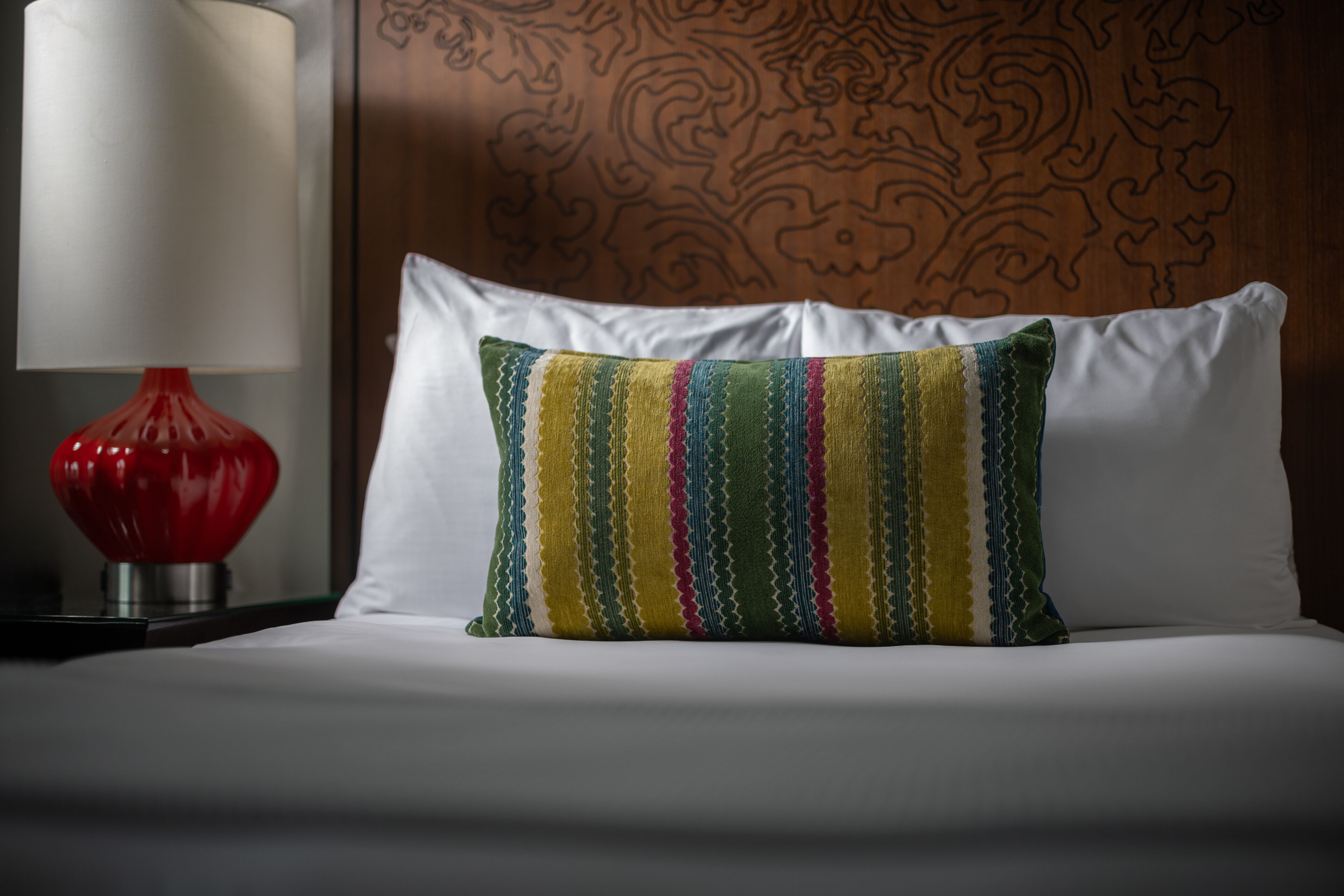 A neatly made bed with a colorful striped pillow, a red lamp, and a patterned wall.