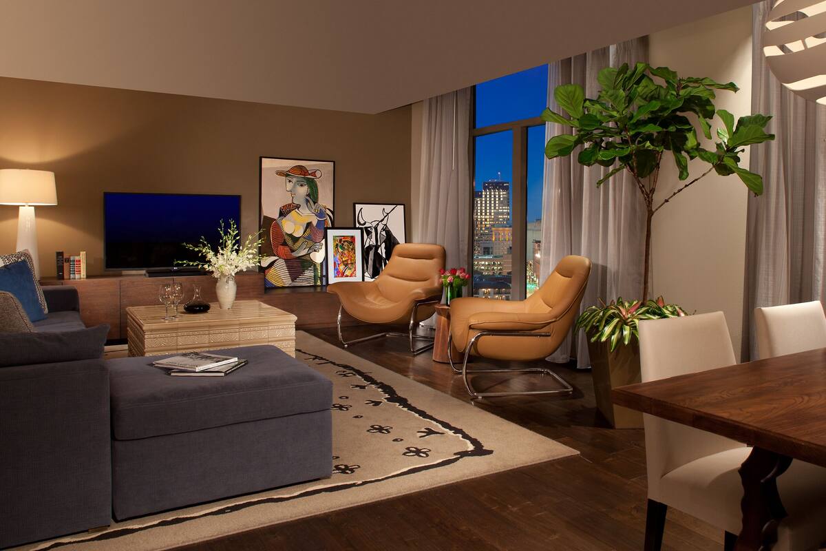 Un salon moderne comprenant un canapé, deux fauteuils, une table basse et une table à manger. Une grande fenêtre offre une vue sur la ville, et on y trouve également un téléviseur à écran plat ainsi que divers objets de décoration, notamment des plantes et des œuvres d’art.