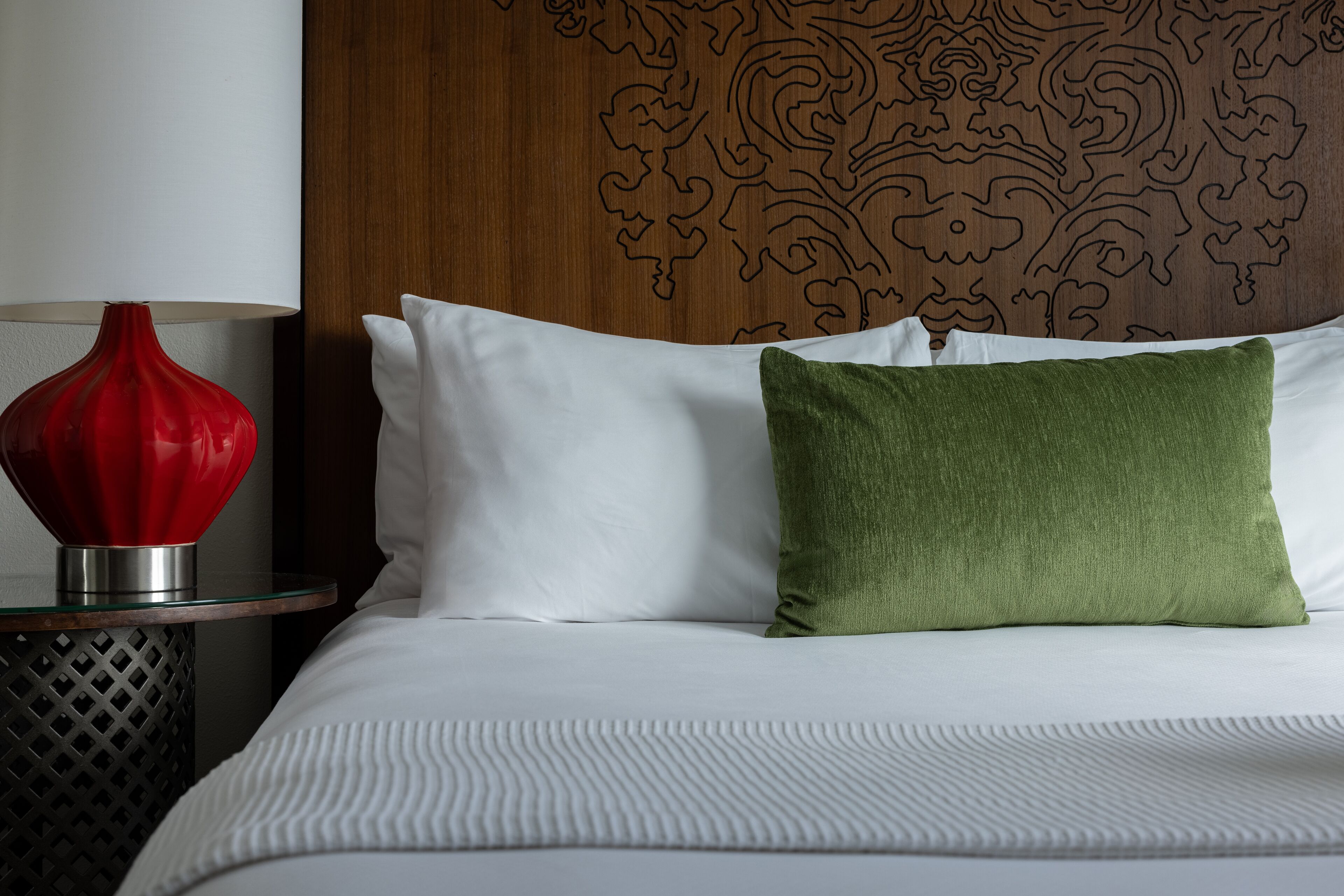 A neatly made bed with white linens and a green pillow, a red vase on a glass table beside it, and a wooden headboard with a carved design.