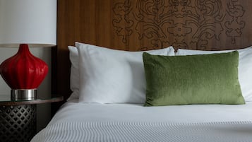 Ein ordentlich bezogenes Bett mit weißen Leinen und einem grünen Kopfkissen, eine rote Vase auf einem Glastisch daneben, und ein Holzkopfteil mit geschnitzten Verzierungen.