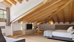 A bedroom with a sloped ceiling, wooden beams, a bed, a desk, a chair, a TV, and a window. - Hotel Venta Etxalar (Etxalar)