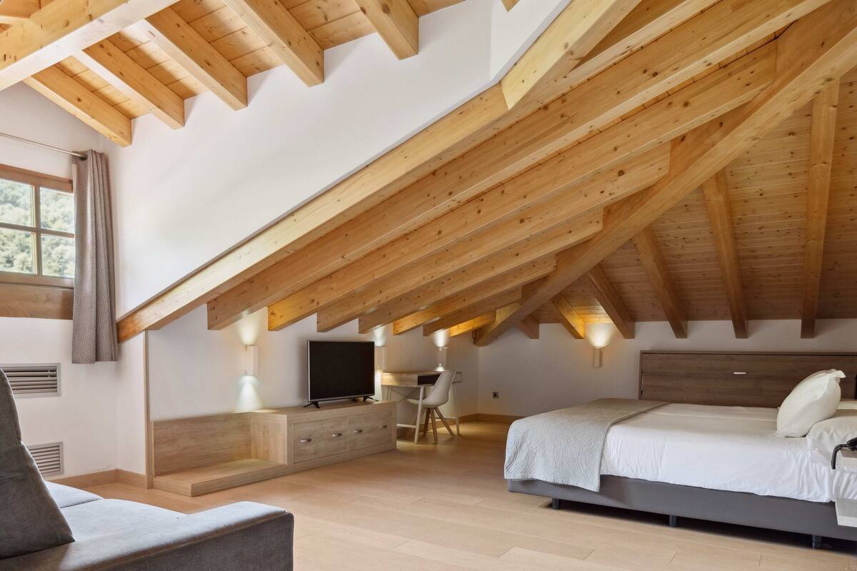 Un dormitorio con techo inclinado, vigas de madera, una cama, un escritorio, una silla, un televisor y una ventana.