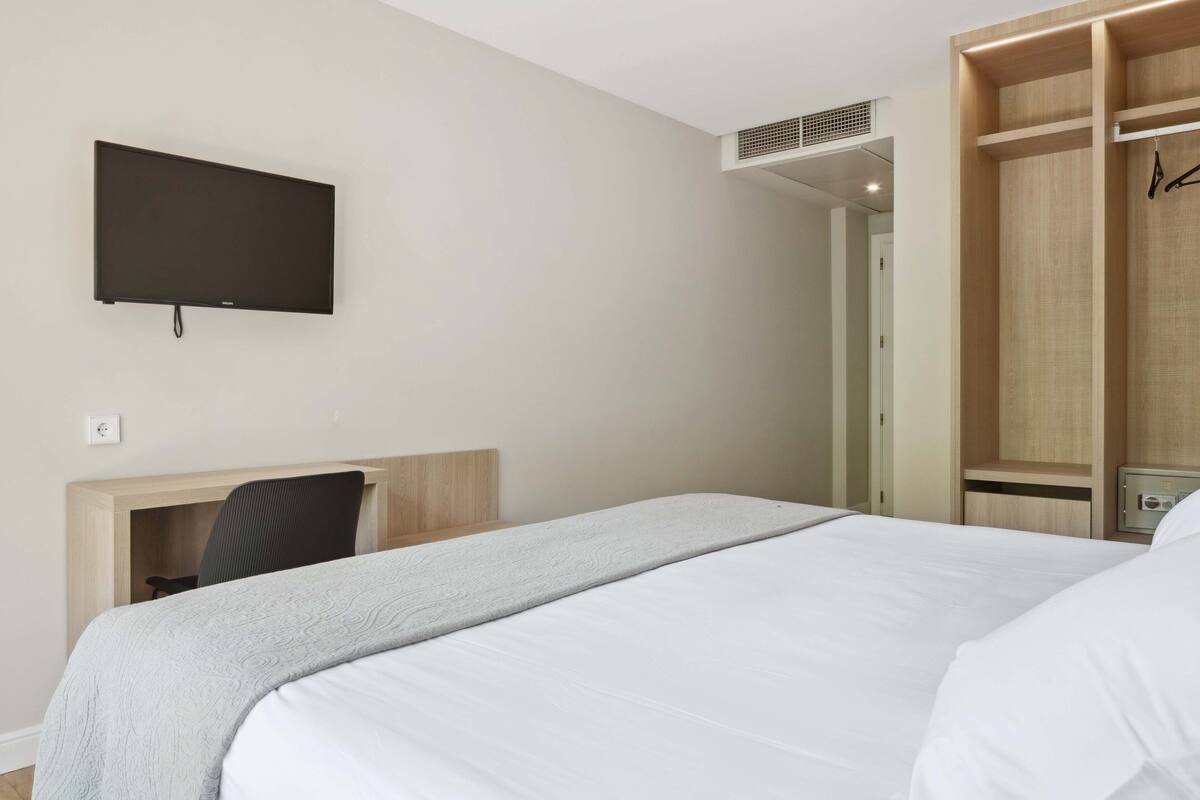 Una habitación de hotel moderna con una cama, un escritorio, una silla, un televisor montado en la pared y un armario.