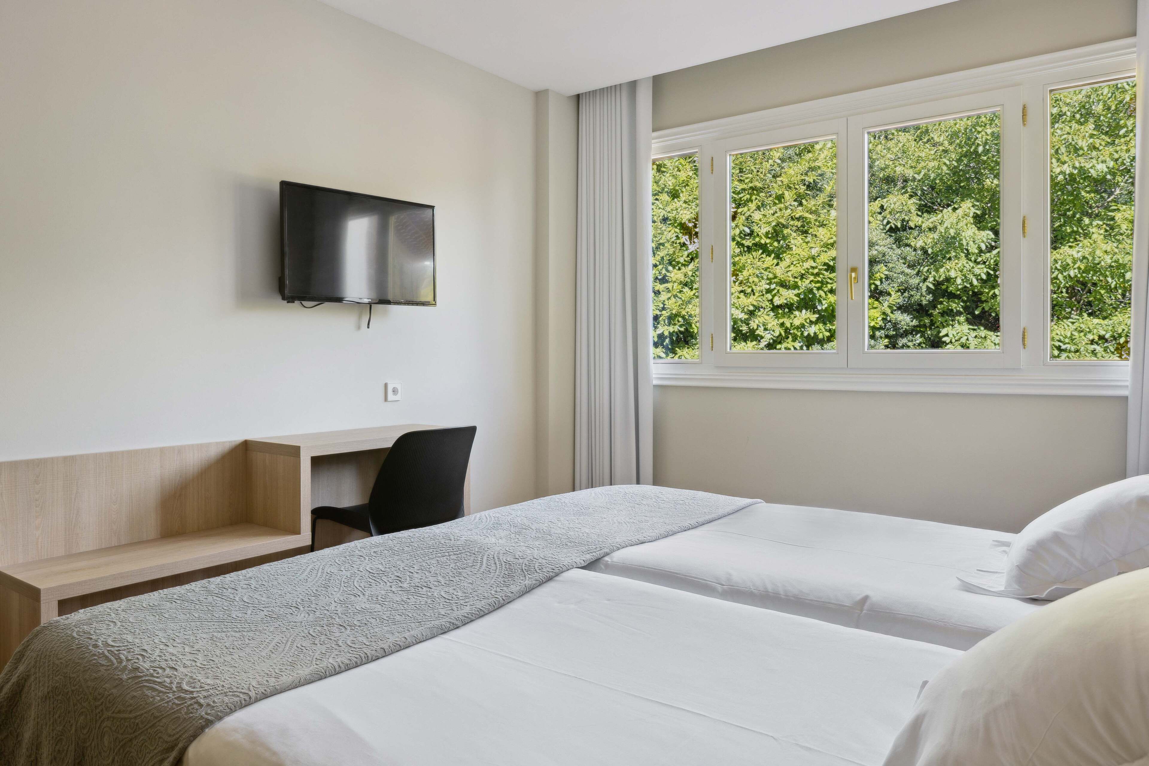 A hotel room with two beds, a TV mounted on the wall, a desk, and a window with a view of trees.
